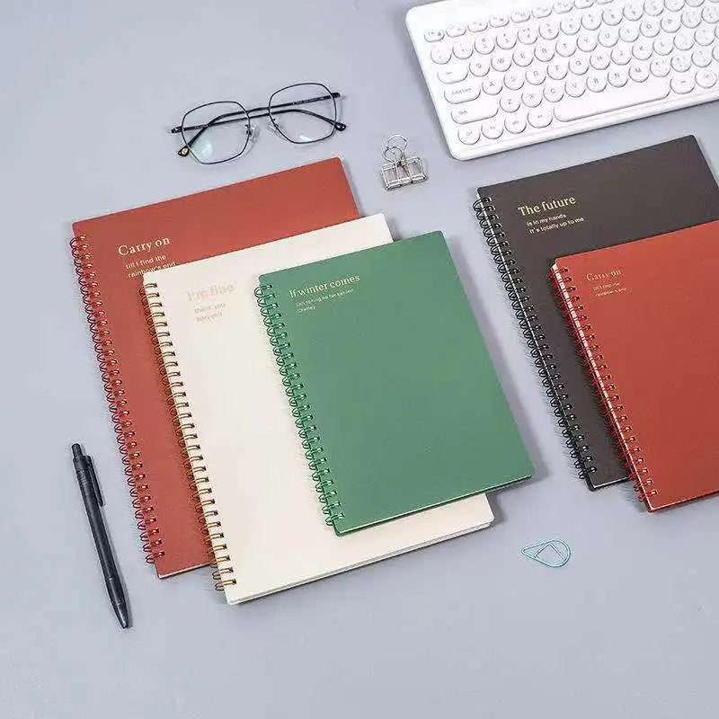 Four spiral notebooks in red, white, green, and brown colors arranged neatly on a work desk, surrounded by office supplies.